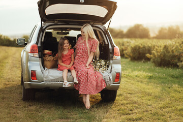 Young Mom with her Little Daughter Dressed Alike in Red Polka Dot Dress, Sitting in a Car Trunk in the Field, Road Trip Picnic Concept