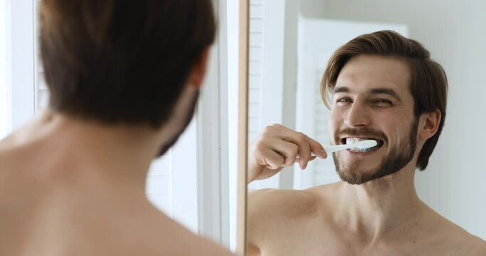 Close up head shot mirror reflection young happy bare man brushing teeth with toothbrush and toothpaste. Smiling handsome guy enjoying morning oral anti cavity hygienic routine alone in bathroom.
