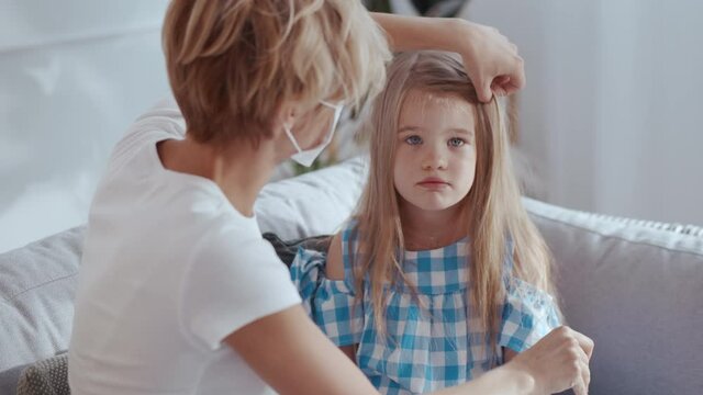 Young Happy Mother Brushing Hair Her Beautiful Blonde Daughter Fixing Hair Clips Preparing For Walking Out. Mom Wearing Protective Mask. Pandemic. Coronavirus.