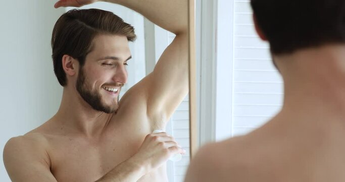 Handsome caucasian man looking in mirror, applying underarm deodorant in arm pit, preventing sweating. Happy young guy using antiperspirant after morning showering, enjoying daily skincare routine.