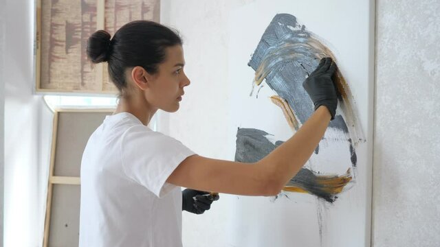 professional girl artist in t-shirt draws painting of black and yellow colours in gloves standing in workshop with white walls in summer