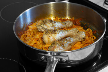 Chicken drumsticks covered in rosemary with tagliatelle pasta and tomato sauce.  Cooking on a hob stove in a frying pan.