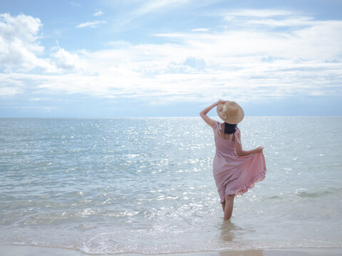 Asian skin tan woman with pink dress and hat she standing relaxing on sand beach with blur image of sea. for travel summer in holidays.