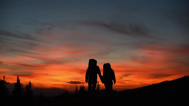 Silhouette of couple in love at sunset in mountains. Guy and girl with backpacks are holding hands on top of mountain under magical sky at sunset, stop and kiss then enjoy view of mountains, forests