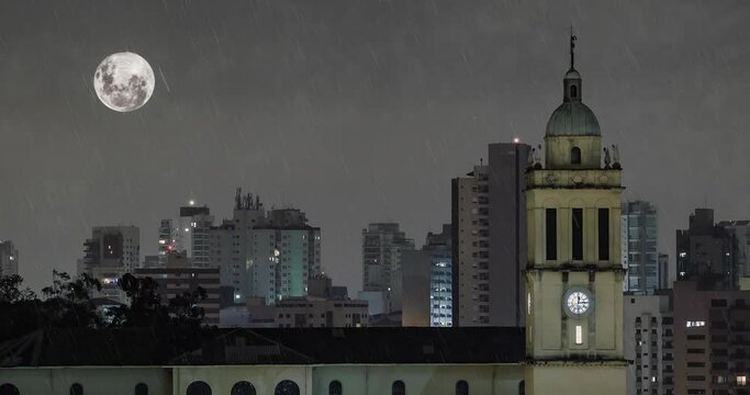 Rainy night of a city during full moon. View to a church tower and the buildings of the city. Animation.