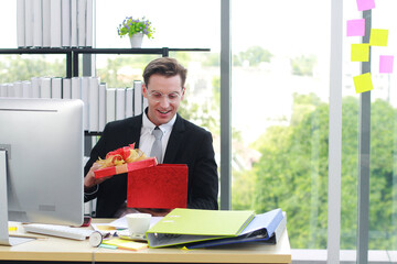 Fototapeta premium Handsome and smart businessman in black suit wear glasses surprise and smiling with red gift box for success in work or happy new year present in Ofiice.Christmas and Happy new year concept
