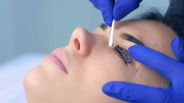 Cosmetologist wiping paint from lashes and brows of woman, side view. Beautician ending lash lifting laminating, painting lashes and eyebrows procedure in cosmetology clinic. Beauty procedure.
