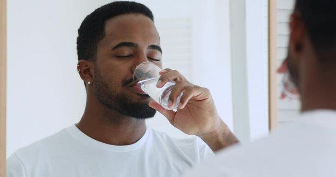 Mirror reflection peaceful young sleepy african american man drinking pure fresh mineral water, waking up in morning. Calm handsome biracial afro ethnic guy doing morning nutrition healthcare routine.