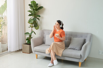 Delighted young woman listening to music with headphones and using mobile phone while leaning on a couch at home