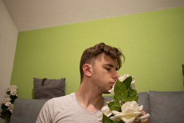 Young male with light brown hair in a white t shirt in a green room smelling a white rose AM