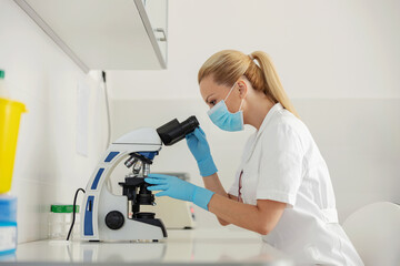 Naklejka na ściany i meble Side view of dedicated blond female lab assistant sitting in laboratory and using microscope for blood test.