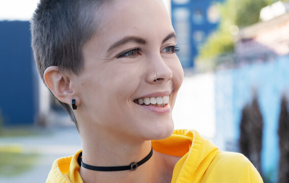 Close up of young smiling woman with shaved hair in urban outdoor city background. Portrait attractive hipster with nose piercing at summer.