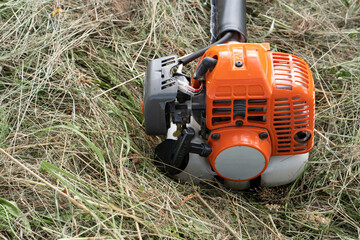 The petrol scythe trimmer brushcutter lies on the hay cut after mowing tall grass in the garden of a country house. Close-up of a petrol brush engine, blurred background, shallow depth of field.