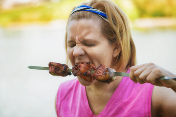 A woman eats meat cooked over an open fire. The summer Bank of the river.