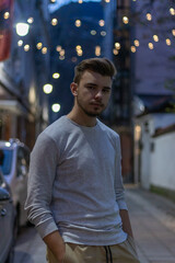 Young male with brown hair in a white long sleeve shirt in an alley with fairy lights installed