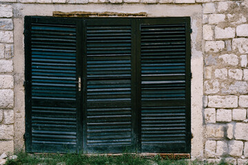 An old closed window in black with a blue tint with blinds on a brick wall.