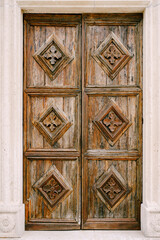 A wooden brown door with carved diamonds and patterns in the opening of a white wall.