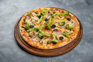 Baked italian pizza with olives, broccoli, ham and mushrooms on a wooden tray on a gray background