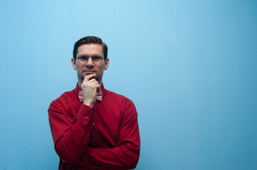 Portrait of thoughtful man in glasses and red shirt posing on blue background.