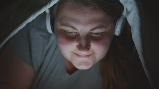 Fat woman at night lying in bed under blanket listening to music in headphones and using smartphone