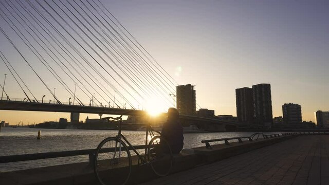 Man with a Bicycle is sitting on the Embarkment near the Erasmus Bridge and smoking joint with marijuana at Sunset time in Rotterdam, Netherlands