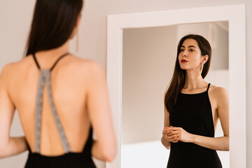 Fototapeta na wymiar Woman in a long black dress with bare back looking at her reflection in the mirror while getting ready for an event, party, date