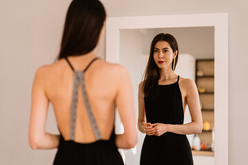 Fototapeta na wymiar Woman in a long black dress with bare back looking at her reflection in the mirror while getting ready for an event, party, date