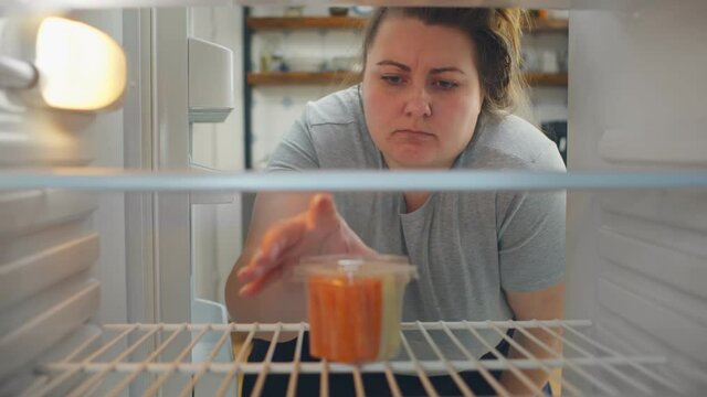 Overweight woman on diet opening empty fridge with only container with vegetables and taking it