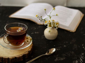 English tea with chamomile stands on a wooden frame and a teaspoon and an open book for reading lies on a black background. Nearby is a white vase with summer chamomile flowers