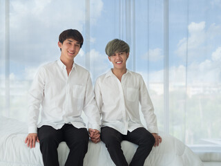 Young Asian LGBT couple sitting on the bed holding hands together with smiling faces, white curtain seeing beautiful blue sky as background in the bedroom. Love, happy together, lifestyle concept.