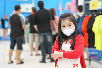 Asian child or kid girl wear white face mask for close mouth nose to flu cough sick for protect PM2.5 dust or coronavirus bacteria to crossed arms on lot of people in department store or shopping mall