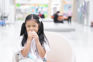 Asian child hungry or kid girl eating bread or holding sandwich before learn at school for morning food or snack breakfast and hanging face mask on neck for coronavirus protect in department store
