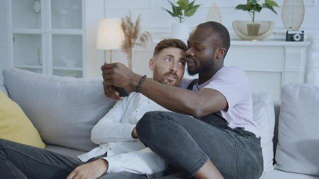 Multi-ethnic affectionate young men gays taking funny selfie pictures on smartphone camera relaxed together in living room. Apartment. LGBT couple.