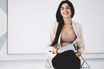 Portrait of cheerful young brunette in trendy eyewear sitting holding modern touchpad on white...