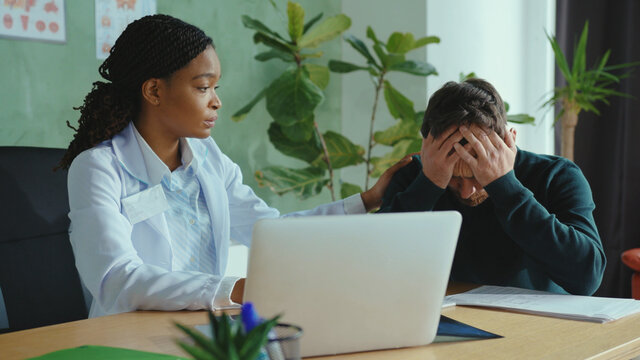 Black Female Doctor Analyzing X-ray Scan Lungs Of Patient Coming Up With Bad Diagnosis. Caring Physician Supporting Young Man And Giving Treatment Instructions.