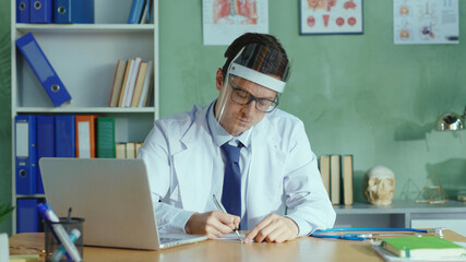 Caucasian doctor wearing protective face shield, writing down medical prescription, looking straight at camera. Concept of coronavirus, health care, quarantine.