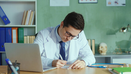 Young professional doctor wearing glasses making notes medical prescription working at table with laptop looking calm confident at camera. Health care workers. Portraits.