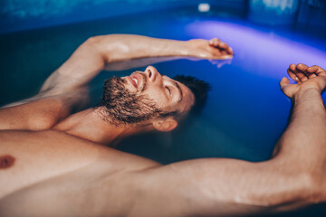 Handsome beard man floating in tank filled with dense salt water used in meditation, therapy, and alternative medicine.