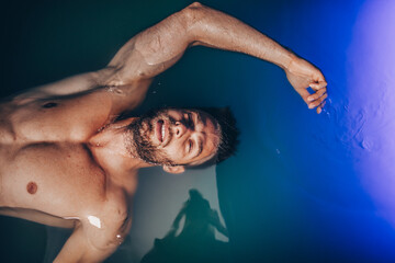 Handsome beard man floating in tank filled with dense salt water used in meditation, therapy, and alternative medicine.