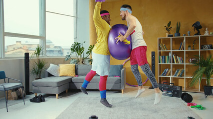 Funny playful unfit young retro styled men playing with fitness ball jumping and fighting in the...