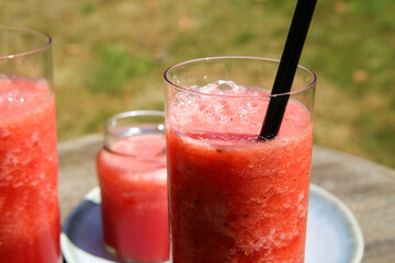 Closeup of isolated ice cold watermelon juice glass with straw outdoor on table. Slices of red fruits on plate.