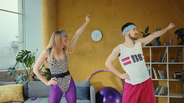 Funny caucasian retro couple doing warm-up aerobics exercises together. Female and male fitness lovers working out training at home in stylish vintage apartment.