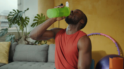 Sweaty and tired african american sportsman drinking water from fitness cup after intensive home workout. Training. Sports activity. Health, nutrition concept.