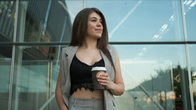 Beautiful caucasian girl with a sincere smile in a gray suit walks near the office and enjoys coffee or tea