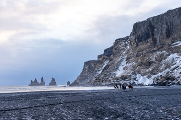 people ride horses on a Vik black beach of the panorama and waves of the winter Atlantic Ocean on Reynisfjara