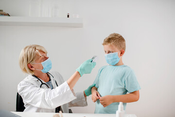 Female doctor doing medical exam to a cute boy. They are wearing a protective face mask.