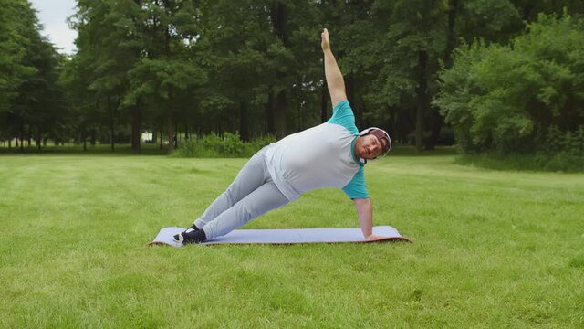 Portrait of stout man in headphones doing yoga exercises on fitness mat outdoors