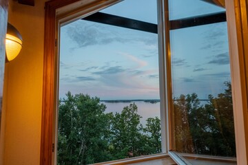 Beautiful summer sundown view from open window. Top of green trees on lake and cloudy sky background. 