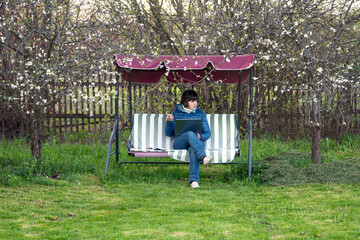 Real female teacher conducts a lesson on the Internet while sitting in the garden on an outdoor garden swing during coronavirus quarantine