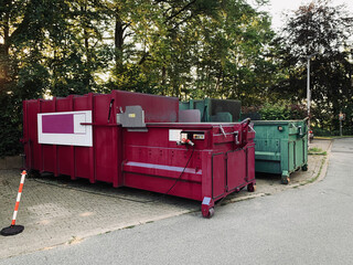on an industrial site a green waste compactor and a red waste compactor stand side by side